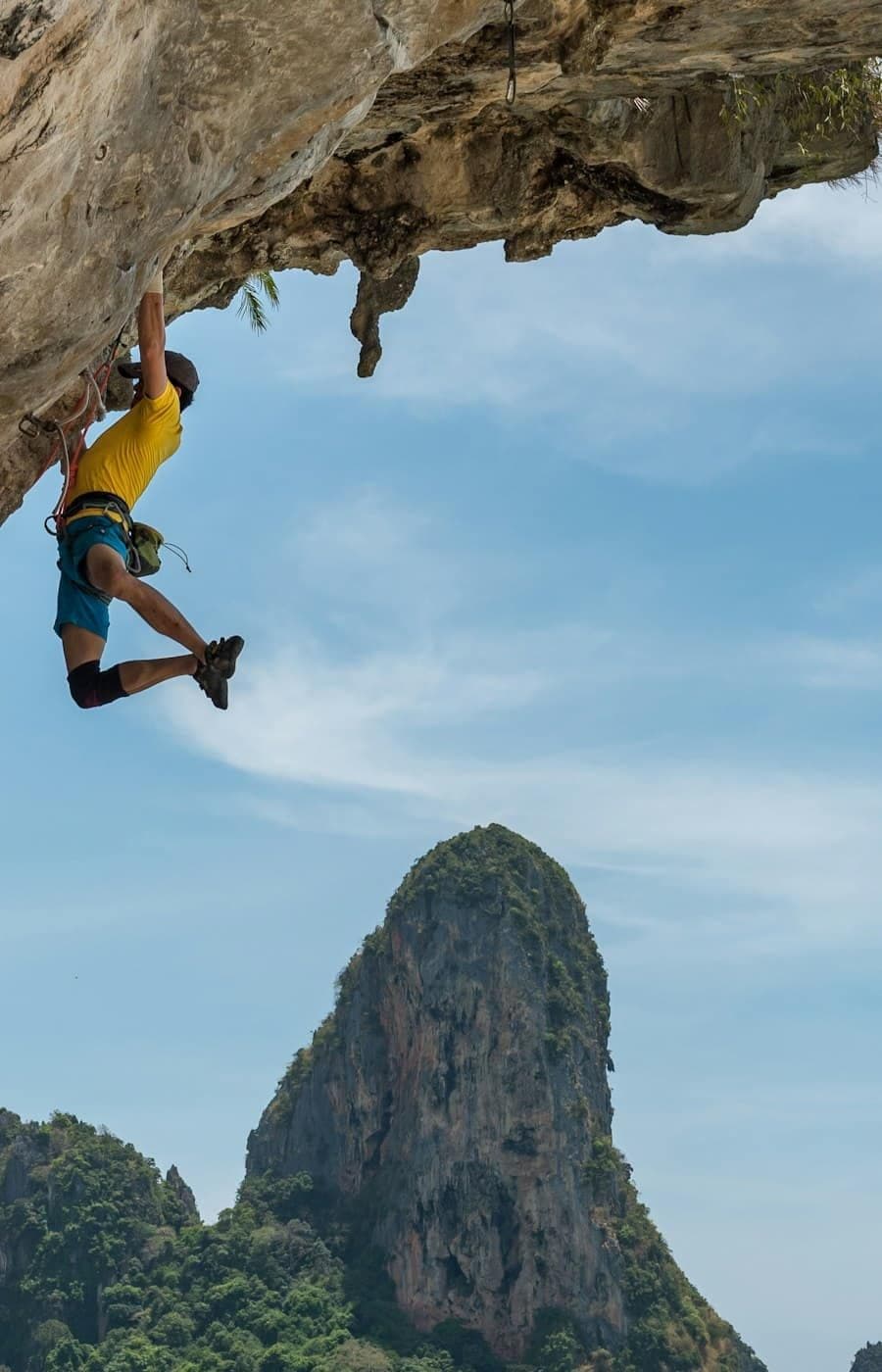 A climber in a yellow shirt hangs from an overhang above a tropical bay.